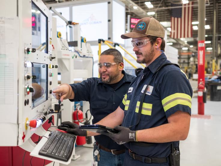 Two men wearing safety gear in a manufacturing setting are looking at computer equipment