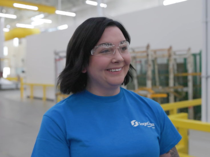 A woman wearing a blue SageGlass shirt and safety goggles walks through a glass factory 