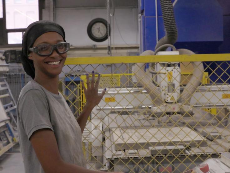 A Black woman wearing safety goggles is standing near a yellow chain link fence inside of a ceramics plant