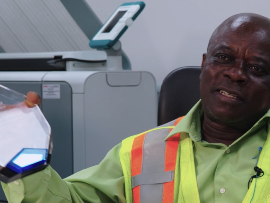 A man wearing a yellow safety vest sits in his office, holding a company award.