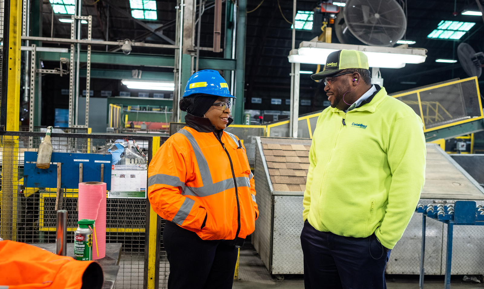 A Black man and a Black woman are standing in a factory setting. She wears a blue hard hat and orange safety jacket. He wears a yellow safety jacket. 