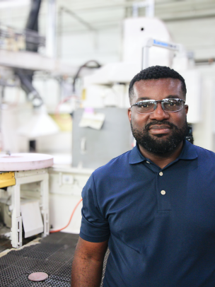 A Black man wearing a blue polo shirt stands in a factory setting in front of white machinery