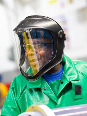 A Black man wearing a green safety suit wears a face shield for his job.